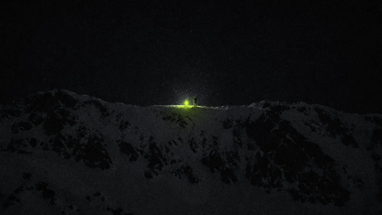 Di notte, sulla cresta di una montagna, una persona minuscola accanto a un fuoco lime: l'unica luce accesa.